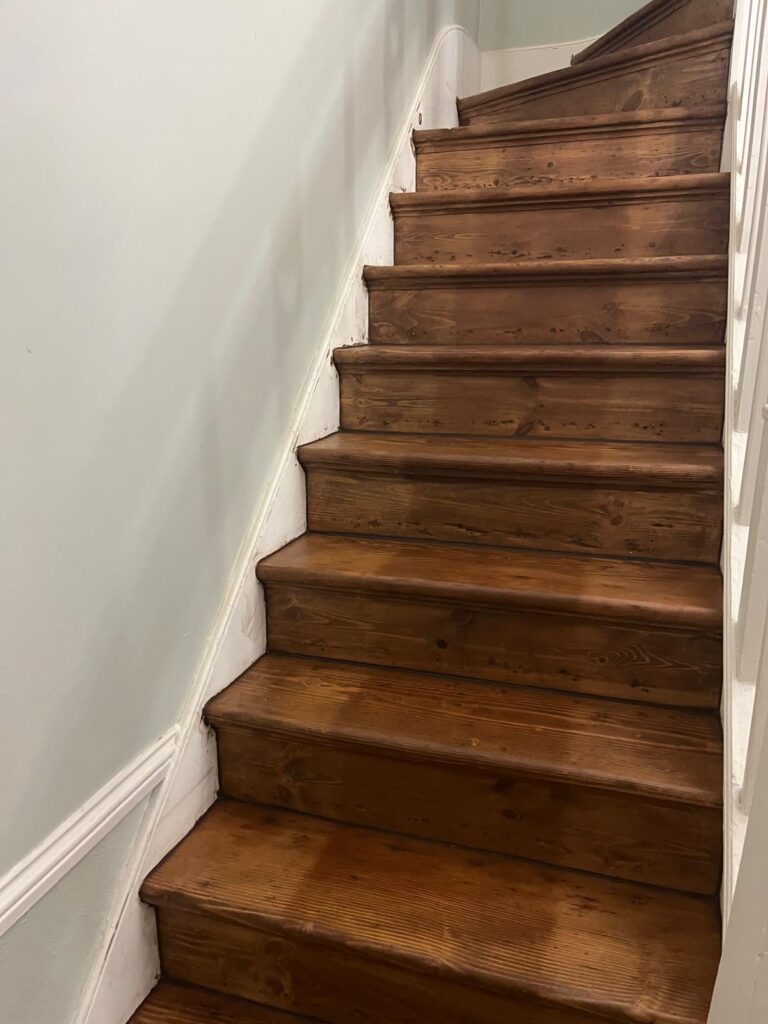 A close-up view of a professional staircase restoration featuring newly stained dark oak wood treads and risers against a light blue wall.