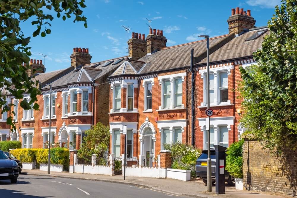 A row of brick houses with white trim and bay windows, lined up along a street with parked cars and a hedge.