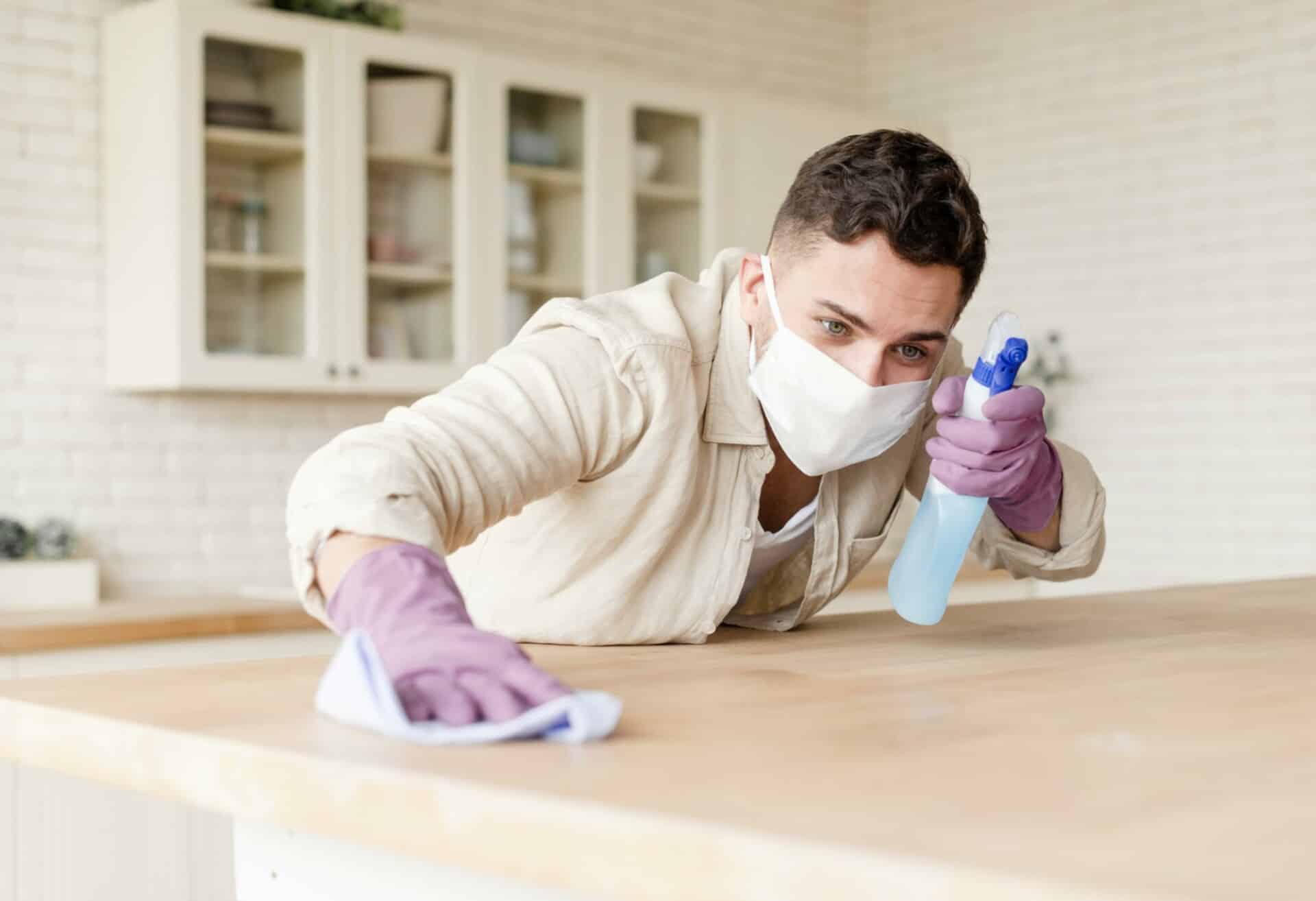 Man in a face mask and gloves cleaning a kitchen counter with a spray bottle and cloth.